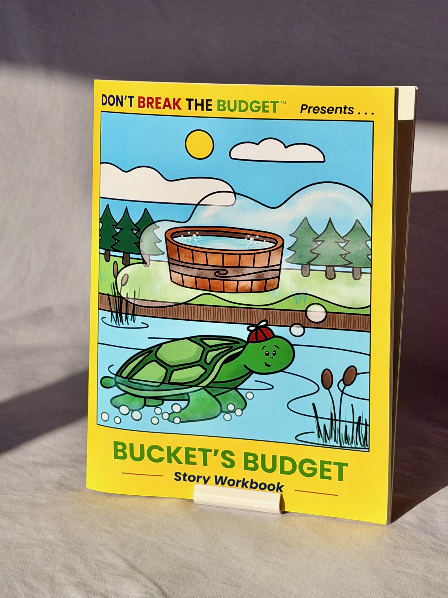 The printed Bucket's Budget story workbook standing upright, showing its bright yellow front cover with Bucket the Turtle swimming beneath a hot tub.