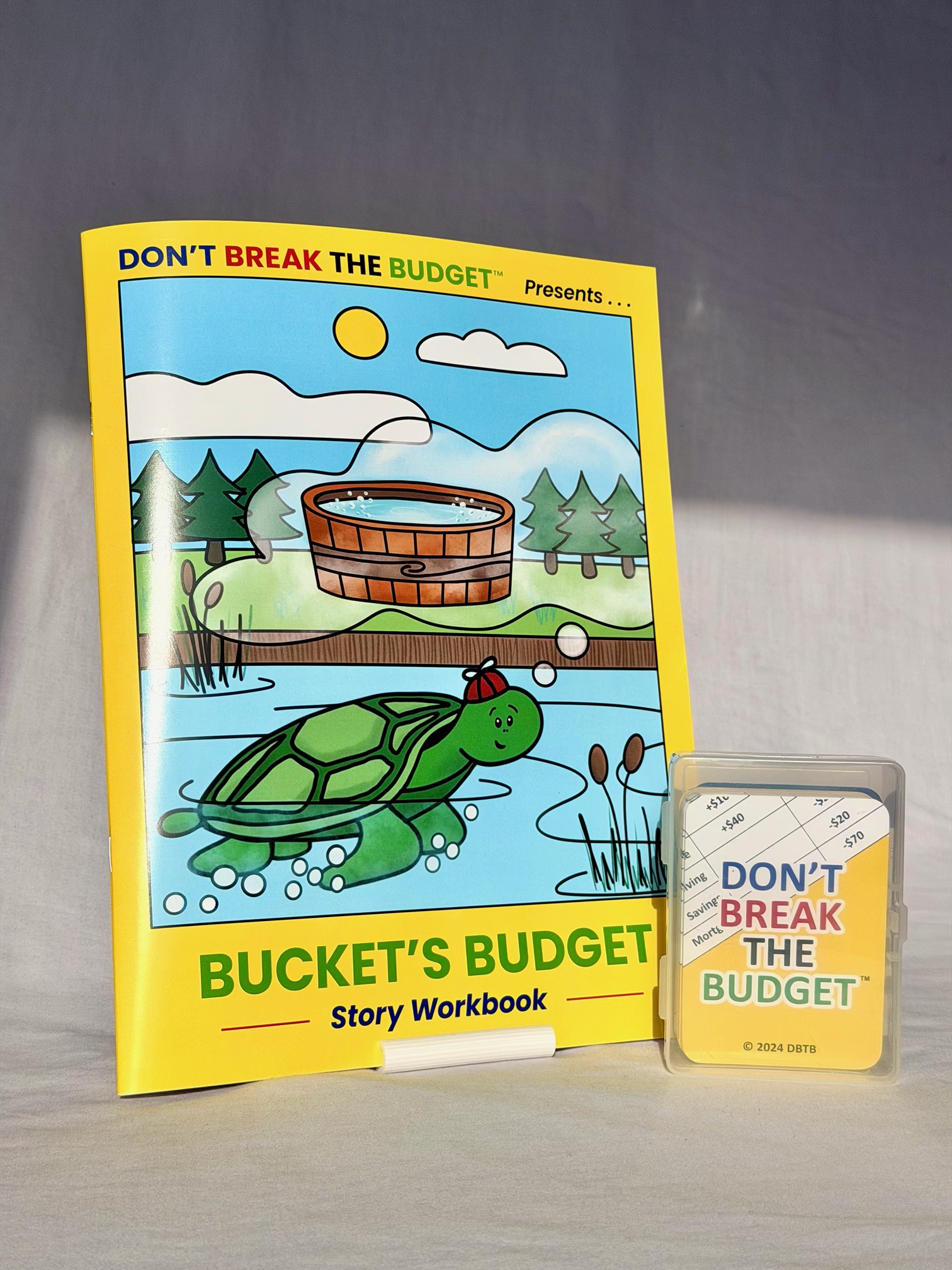The printed Bucket's Budget story workbook standing upright next to the Don't Break the Budget card deck in its clear case.
